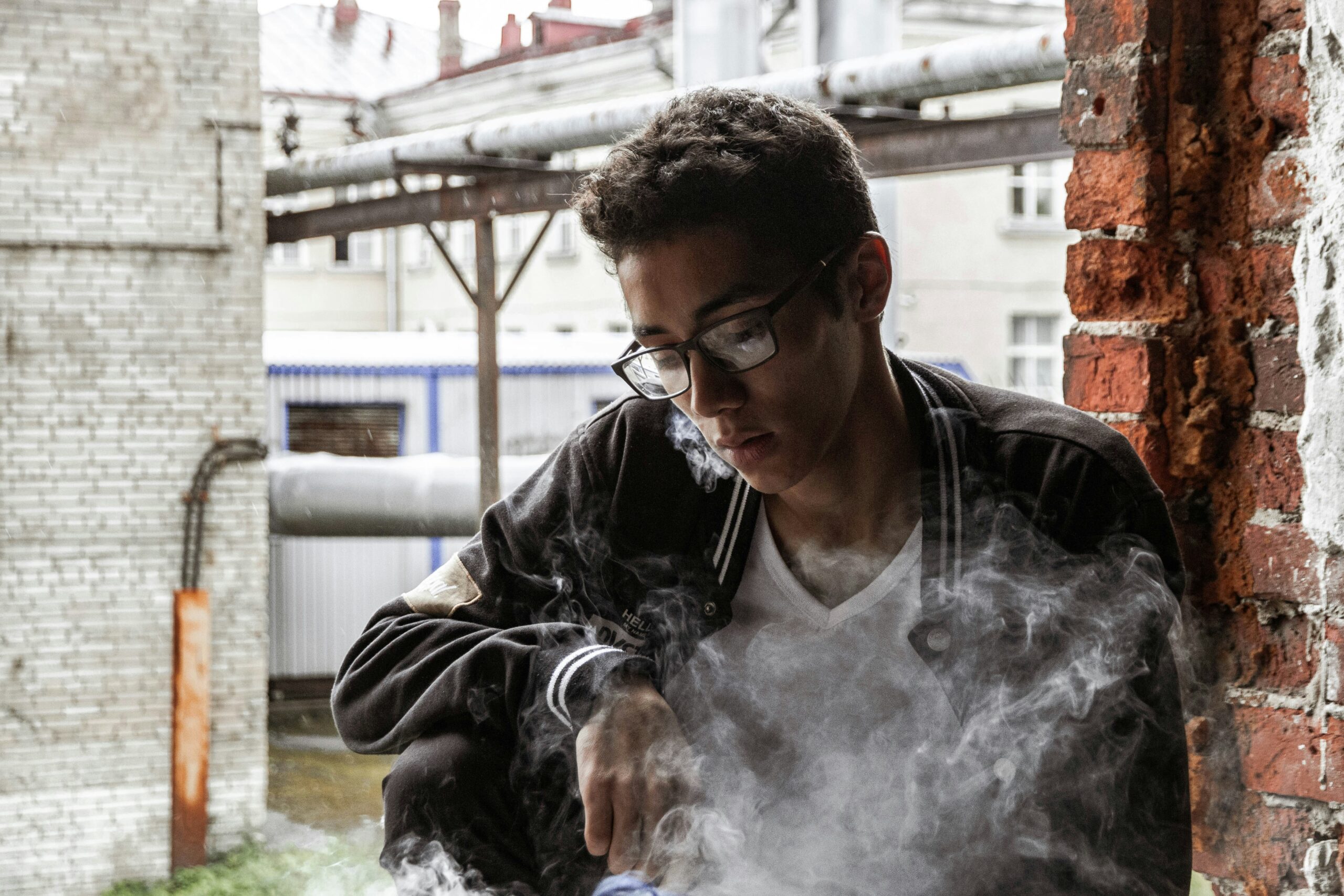 Teenager vaping while sitting in an industrial-looking outdoor space, surrounded by a cloud of vapor.
